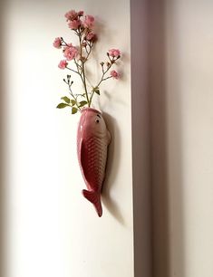 a fish shaped vase with pink flowers hanging on the wall next to it's side