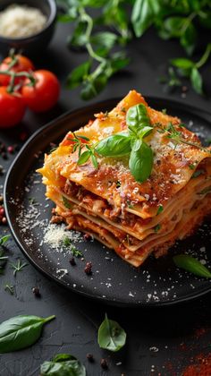 a black plate topped with lasagna covered in cheese and basil next to tomatoes