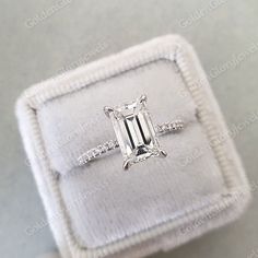 an emerald cut diamond ring in a white velvet box with diamonds on the band and side stones