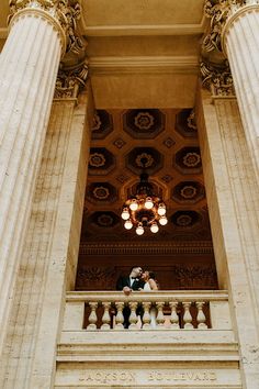 a bride and groom standing on the balcony of an ornate building with columns, chandelier and lights