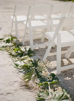 white chairs lined up on the beach with flowers and greenery lining the aisle for an outdoor ceremony