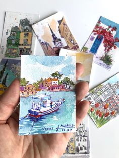 a person holding up some cards with watercolors on them and buildings in the background