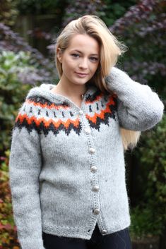 a woman wearing a gray sweater with an orange and black design on the front, standing in front of some bushes