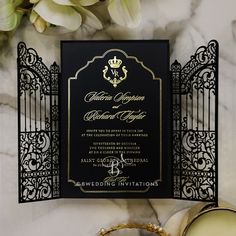 an ornate black and gold wedding card on top of a marble table next to flowers