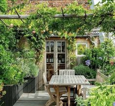 an outdoor dining table surrounded by greenery and potted plants in front of a brick building