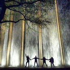 three people holding hands while standing in front of a water fountain at night with their arms around each other