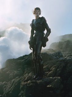 a woman standing on top of a rocky hill next to a cloud filled sky and clouds