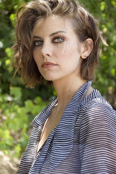 a woman with brown hair and blue shirt posing for a photo in front of trees