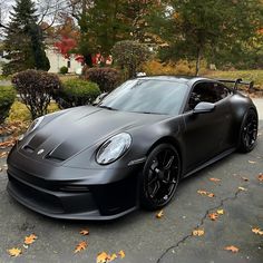 a black sports car parked in front of some bushes and trees with orange leaves on the ground