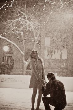 a man kneeling down next to a woman under an umbrella on a snowy day in the city