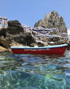 a red boat floating on top of a body of water next to rocks and umbrellas