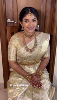 a woman in a white and gold sari sitting on the floor with her hands together