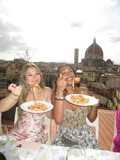 two women eating pasta on a rooftop in rome, with the city skyline in the background