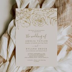 a white and gold wedding card on top of some fabric with flowers in the middle