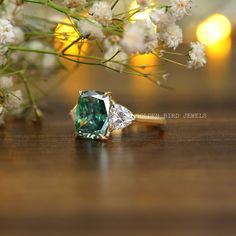 a green diamond ring sitting on top of a wooden table next to white flowers and greenery