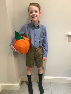 a young boy in overalls holding an orange pumpkin and smiling at the camera while standing next to a wall