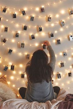 a woman is sitting on her bed with lights strung over the headboard and pictures hanging from the wall