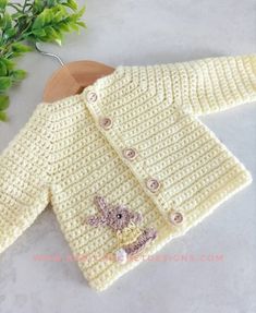 a crocheted sweater with buttons and a teddy bear on the front is next to a plant