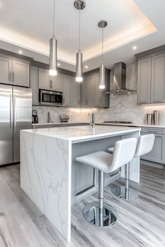 Modern kitchen with grey cabinets, stainless steel appliances, marble countertops, and two bar stools at an island. White Modern Kitchen Apartment, White Kitchen Grey Cabinets, White And Silver Kitchen Ideas, White Grey Gold Kitchen, Kitchen Ideas White And Grey, Kitchen Design White And Grey, White And Grey Kitchen Decor, Kitchen Design Grey And White, White And Gray Kitchen Decor