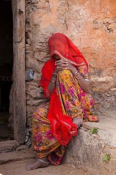 a woman sitting on the ground in front of a stone building wearing a red head scarf