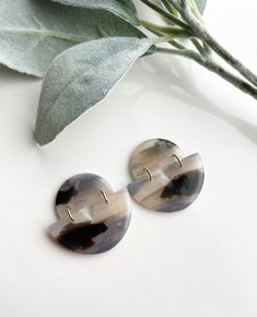 two round shaped earrings sitting on top of a white surface next to a plant with green leaves