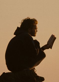 a man sitting on top of a rock while reading a book in the sky at sunset