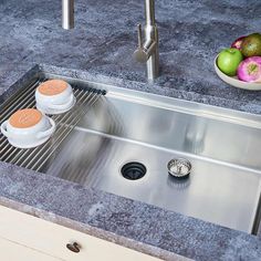 a stainless steel kitchen sink with two cups on the drain and some fruit in the background