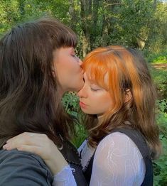 two women kissing each other in front of trees and bushes, with one woman's face to the side