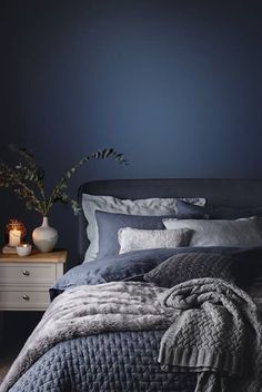 a bedroom with dark blue walls and grey bedding in the foreground is a white table with two vases on it