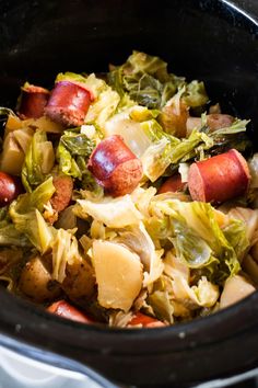 a crock pot filled with cabbage, potatoes and hot dogs on top of a stove