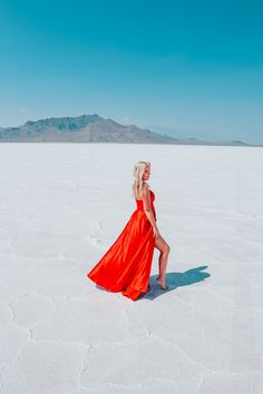 a woman in a long red dress walking across a salt flat area with mountains in the background