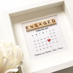 a wooden scrabbled calendar with the word engaged on it in front of a white flower