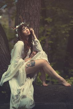 a woman sitting on top of a tree trunk wearing a white dress and flower crown