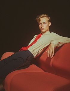 a young man sitting on top of a red couch wearing a shirt and tie with his legs crossed