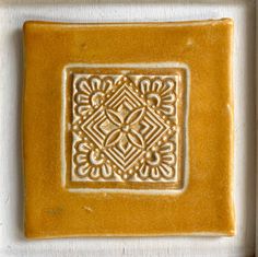 a square tile with an intricate design on the bottom in yellow and white, is shown