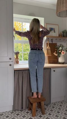 a woman standing on a stool in front of a kitchen window looking out the window