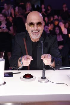 a bald man sitting at a table in front of a microphone and cell phone with people behind him