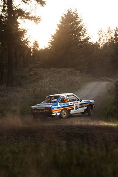 a rally car driving down a dirt road in the woods at sunset or sunrise time