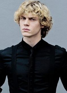 a young man with curly hair wearing a black shirt and tie, standing in front of a gray background