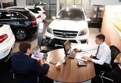 two men sitting at a table in front of a car dealership with their laptops