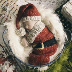 a baby wearing a santa suit and hat sleeping in a bowl with presents on the floor