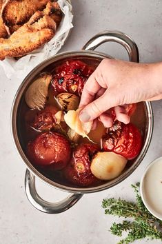 A hand holds a peeled garlic clove above a pot filled with roasted tomatoes and onions, perfect for making Roasted Tomato Soup. Nearby, a plate with toasted bread and a sprig of thyme adds a touch of elegance. Best Roasted Tomato Soup, Fire Roasted Tomato Soup, Immersion Blender Recipes, Roast Tomato Soup Recipe, Blender Soup, Fresh Tomato Soup, Oven Roasted Tomatoes, Slow Roasted Tomatoes, Tomato Soup Homemade