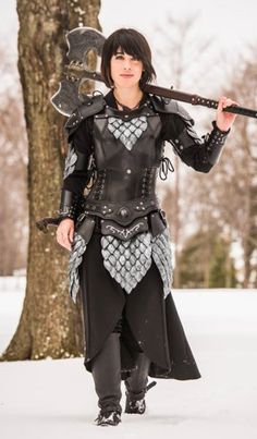 a woman dressed in black and silver holding an ax on her shoulder while walking through the snow