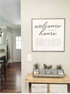 a welcome sign hanging on the wall above a wooden table with two planters in front of it