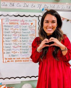 a woman in a red dress making a heart shape with her hands while standing next to a whiteboard