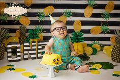 a baby sitting on a table with pineapples in front of him and wearing a party hat