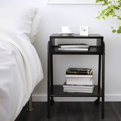 a black end table with some books on it next to a white bed and plant