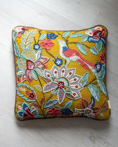 a yellow pillow with colorful flowers and birds on the front, sitting on a white wooden floor