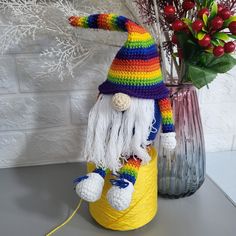 a colorful crocheted gnome sitting on top of a yellow barrel next to a vase with flowers