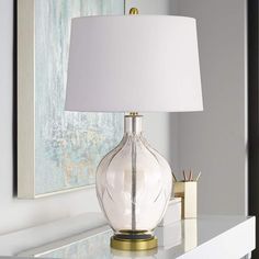 a clear glass lamp sitting on top of a white table next to a framed painting